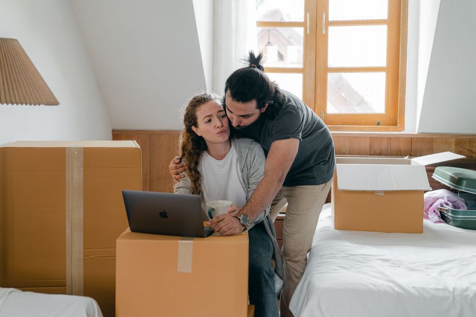 A man and a woman sit close together at a wooden table in a well-lit room filled with packing and moving supplies. The man, wearing a grey hoodie, is holding a pair of shoes in his hands, examining th