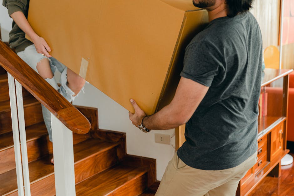 A person is seen inside a well-lit room, likely a living area, preparing for house removal by sealing a large cardboard moving box with a handheld tape dispenser. Several other packed cardboard boxes