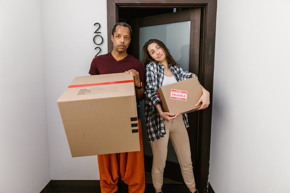 A man and a woman stand inside a narrow, well-lit interior space, near a doorway with a dark wooden frame, during a home relocation process. The man, dressed in a maroon t-shirt and orange trousers, h