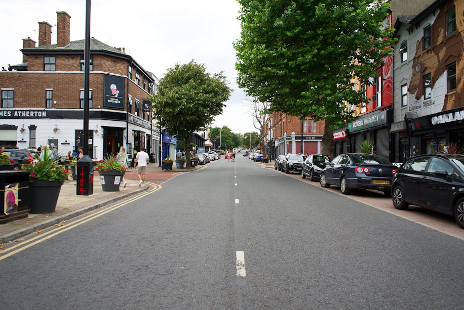 A daytime street scene in Hounslow with a row of multi-storey residential buildings featuring brick facades, gabled roofs, and large windows. The ground level includes various shopfronts, one of which